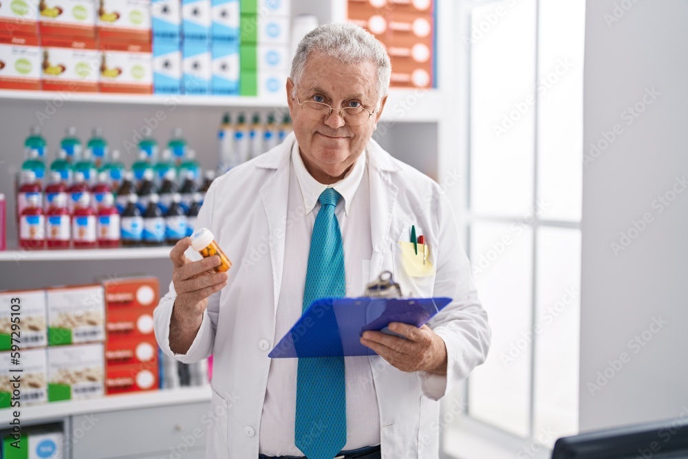 Middle age grey-haired man pharmacist holding pills bottle reading document at pharmacy Middle age grey-haired man pharmacist holding pills bottle reading document at pharmacy