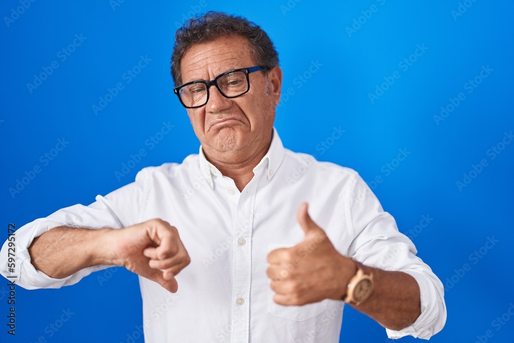 Middle age hispanic man standing over blue background doing thumbs up and down, disagreement and agreement expression. crazy conflict Middle age hispanic man standing over blue background doing thumbs up and down, disagreement and agreement expression. crazy conflict