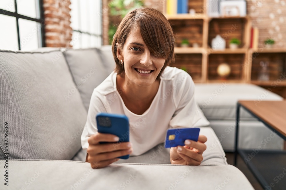 Young beautiful hispanic woman using smartphone and credit card lying on sofa at home Young beautiful hispanic woman using smartphone and credit card lying on sofa at home