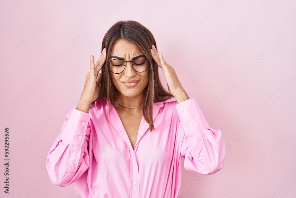 Young hispanic woman wearing glasses standing over pink background with hand on head, headache because stress. suffering migraine.