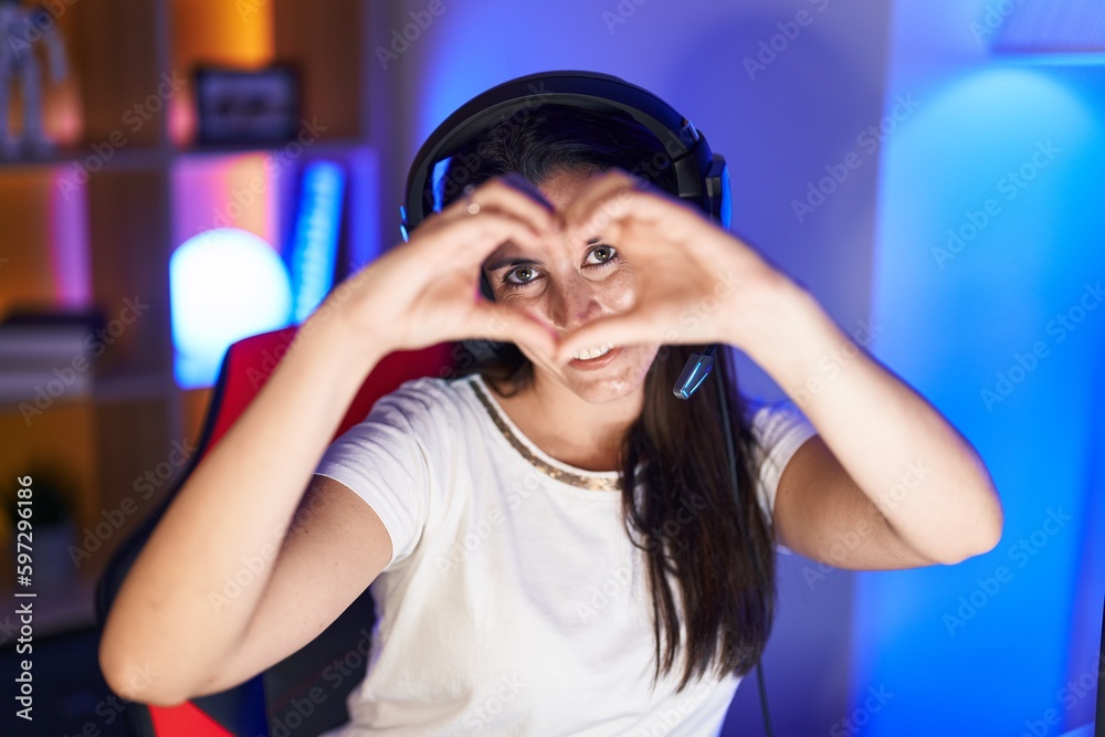 Young beautiful hispanic woman streamer smiling confident doing heart symbol with hands at gaming room