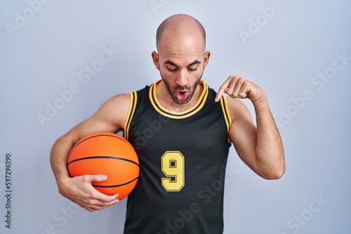 Young bald man with beard wearing basketball uniform holding ball pointing down with fingers showing advertisement, surprised face and open mouth