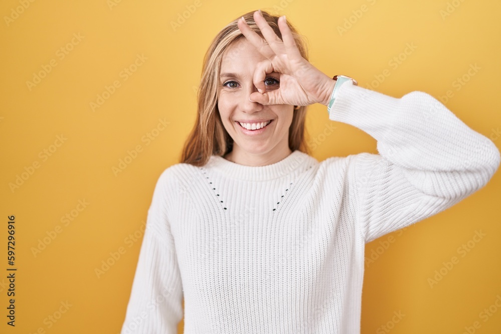 Young caucasian woman wearing white sweater over yellow background doing ok gesture with hand smiling, eye looking through fingers with happy face. Young caucasian woman wearing white sweater over yellow background doing ok gesture with hand smiling, eye looking through fingers with happy face.