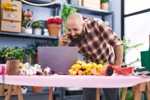 Young bald man florist talking on smartphone using laptop at flower shop