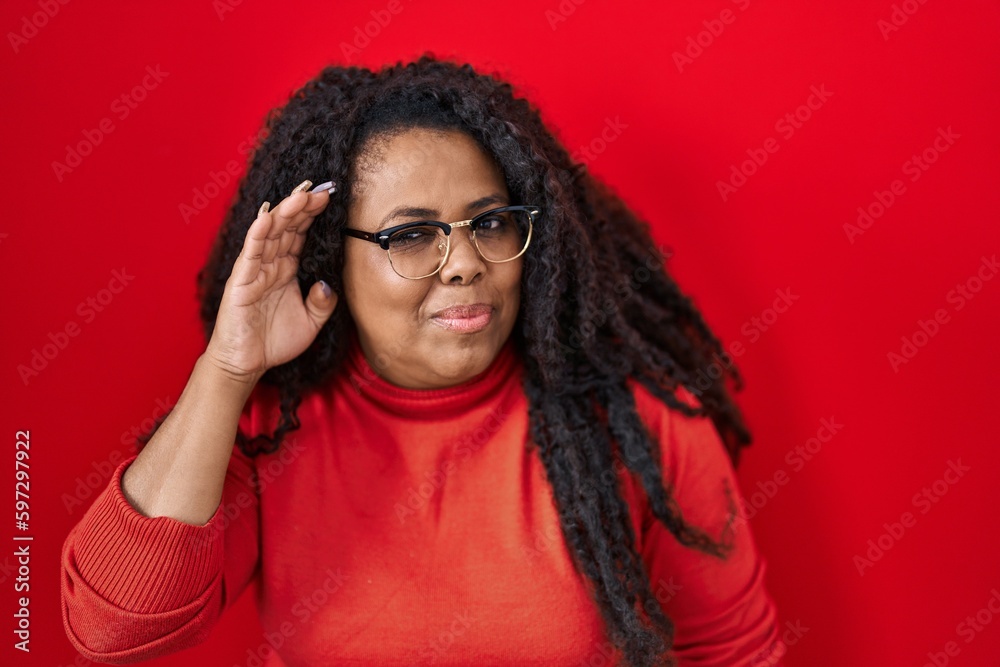 Plus size hispanic woman standing over red background smiling with hand over ear listening an hearing to rumor or gossip. deafness concept. Plus size hispanic woman standing over red background smiling with hand over ear listening an hearing to rumor or gossip. deafness concept.