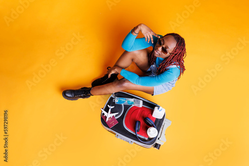 African american girl with sunglasses, posing in studio on the floor with suitcase and items. Cheerful person having trolley bag with multiple essentials and objects, travelling on vacation.