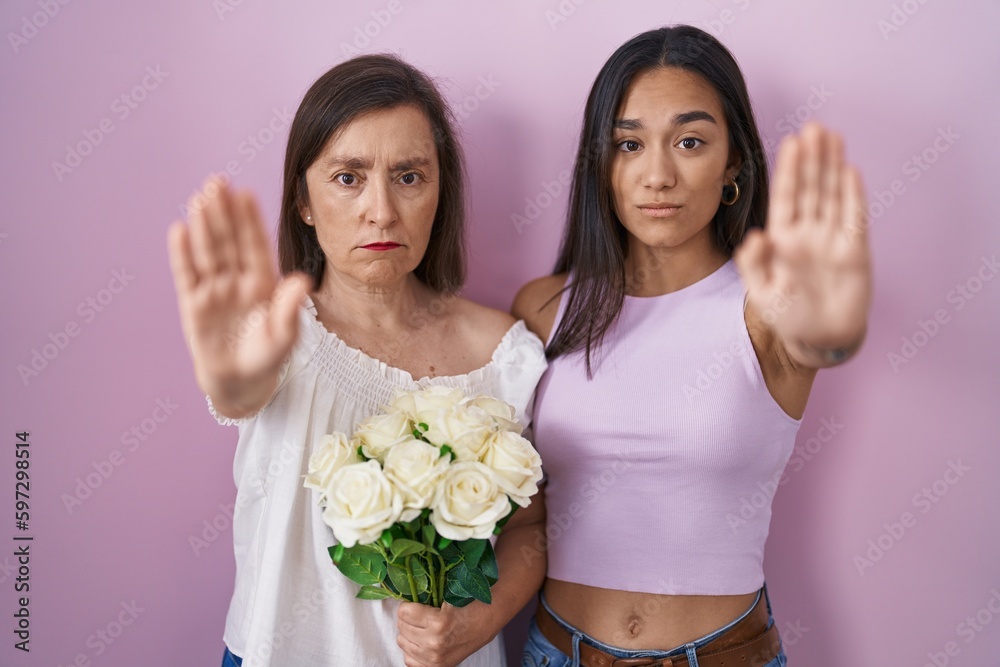 Hispanic mother and daughter holding bouquet of white flowers doing stop sing with palm of the hand. warning expression with negative and serious gesture on the face. Hispanic mother and daughter holding bouquet of white flowers doing stop sing with palm of the hand. warning expression with negative and serious gesture on the face.