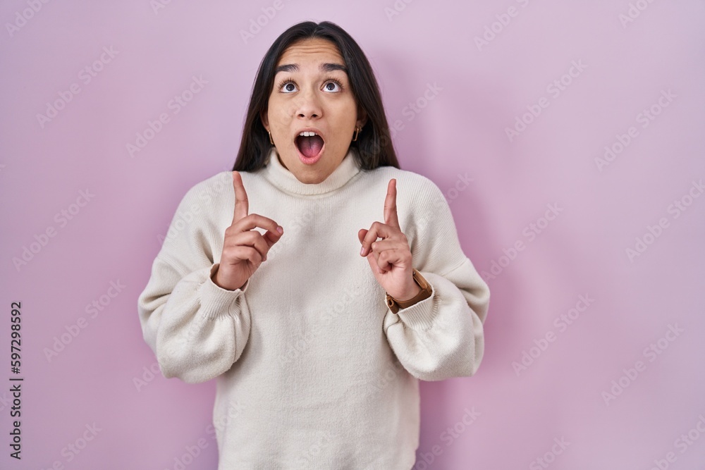 Young south asian woman standing over pink background amazed and surprised looking up and pointing with fingers and raised arms. Young south asian woman standing over pink background amazed and surprised looking up and pointing with fingers and raised arms.