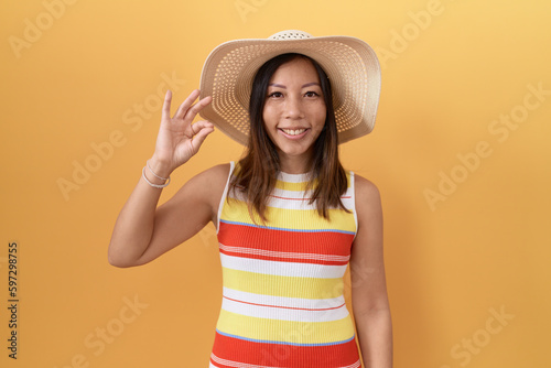 Middle age chinese woman wearing summer hat over yellow background smiling positive doing ok sign with hand and fingers. successful expression.