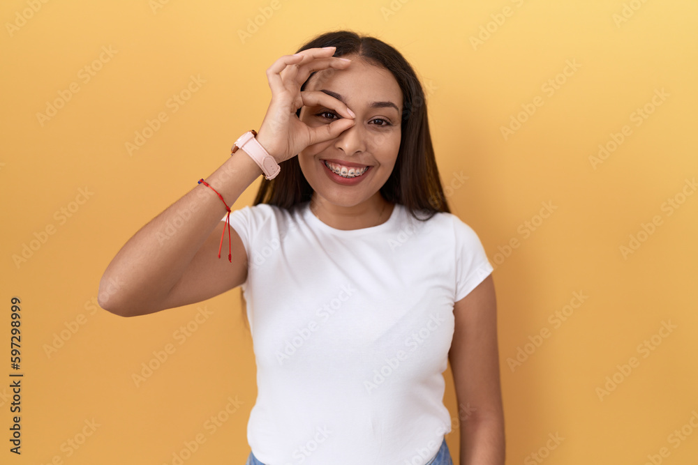 Young arab woman wearing casual white t shirt over yellow background doing ok gesture with hand smiling, eye looking through fingers with happy face. Young arab woman wearing casual white t shirt over yellow background doing ok gesture with hand smiling, eye looking through fingers with happy face.