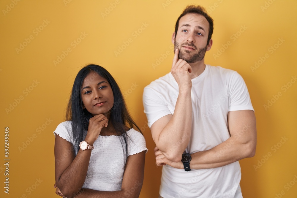 Interracial couple standing over yellow background with hand on chin thinking about question, pensive expression. smiling with thoughtful face. doubt concept. Interracial couple standing over yellow background with hand on chin thinking about question, pensive expression. smiling with thoughtful face. doubt concept.