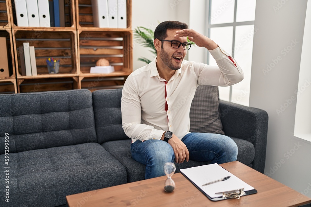 Young hispanic man with beard working at consultation office very happy and smiling looking far away with hand over head. searching concept. Young hispanic man with beard working at consultation office very happy and smiling looking far away with hand over head. searching concept.