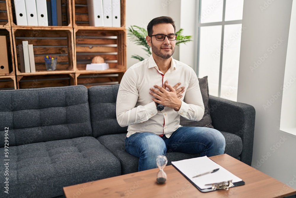 Young hispanic man with beard working at consultation office smiling with hands on chest with closed eyes and grateful gesture on face. health concept. Young hispanic man with beard working at consultation office smiling with hands on chest with closed eyes and grateful gesture on face. health concept.