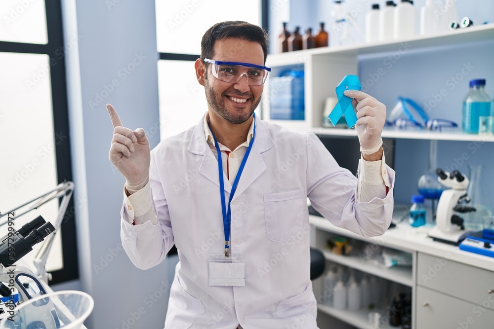 Young hispanic man with beard working at scientist laboratory holding blue ribbon smiling happy pointing with hand and finger to the side Young hispanic man with beard working at scientist laboratory holding blue ribbon smiling happy pointing with hand and finger to the side