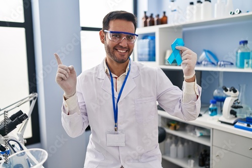 Young hispanic man with beard working at scientist laboratory holding blue ribbon smiling happy pointing with hand and finger to the side