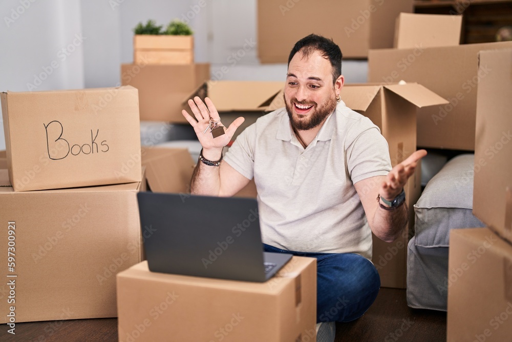 Plus size hispanic man with beard moving to a new home doing video call celebrating achievement with happy smile and winner expression with raised hand Plus size hispanic man with beard moving to a new home doing video call celebrating achievement with happy smile and winner expression with raised hand
