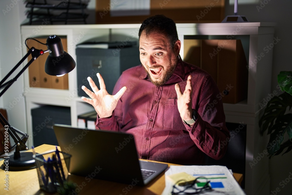 Plus size hispanic man with beard working at the office at night celebrating crazy and amazed for success with arms raised and open eyes screaming excited. winner concept Plus size hispanic man with beard working at the office at night celebrating crazy and amazed for success with arms raised and open eyes screaming excited. winner concept