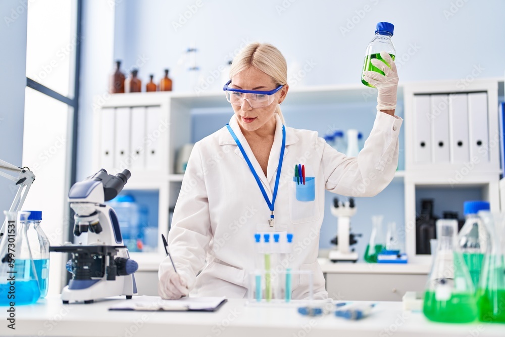 Young blonde woman wearing scientist uniform holding bottle at laboratory Young blonde woman wearing scientist uniform holding bottle at laboratory