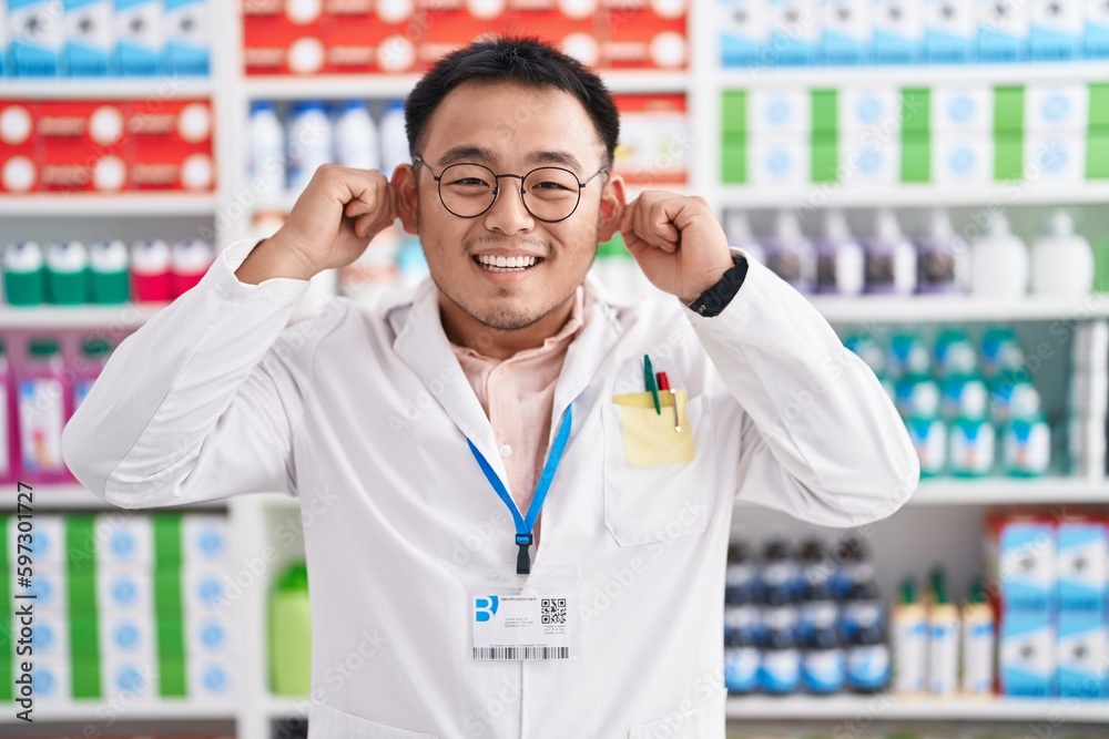 Chinese young man working at pharmacy drugstore smiling pulling ears with fingers, funny gesture. audition problem