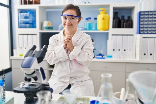 Hispanic girl with down syndrome working at scientist laboratory hands together and fingers crossed smiling relaxed and cheerful. success and optimistic