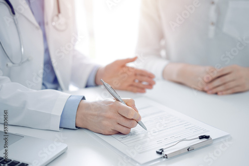 Doctor and patient discussing current health questions while sitting near of each other and using clipboard at the table in clinic, just hands closeup. Medicine concept