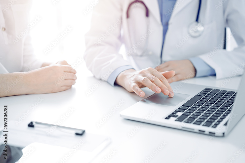 Doctor and patient sitting near each other at the desk in clinic. The focus is on female physician's hands using laptop computer, close up. Medicine concept Doctor and patient sitting near each other at the desk in clinic. The focus is on female physician's hands using laptop computer, close up. Medicine concept