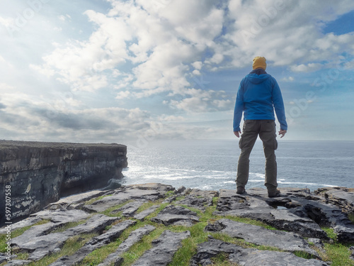 Man tourist in yellow hat and blue jacket standing on an edge of a cliff by the ocean. Aran island, county Galway, Ireland. Stunning nature scenery in the background with ocean and blue cloudy sky