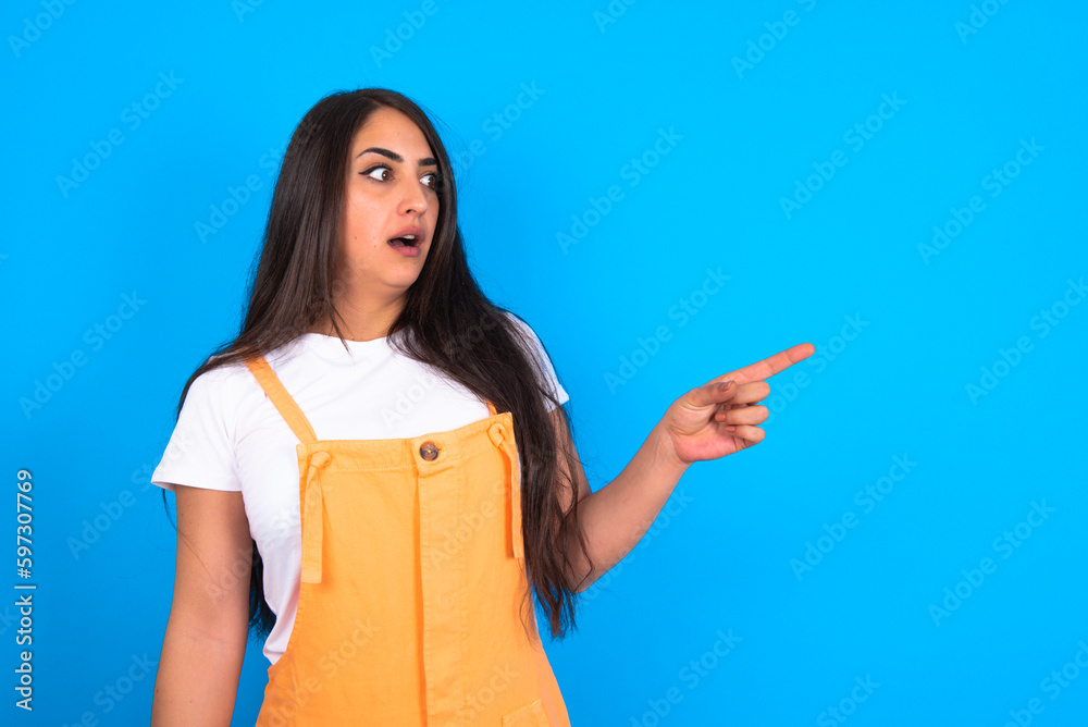 Stunned beautiful brunette woman wearing overall over blue background with greatly surprised expression points away on copy space, indicates something Stunned beautiful brunette woman wearing overall over blue background with greatly surprised expression points away on copy space, indicates something