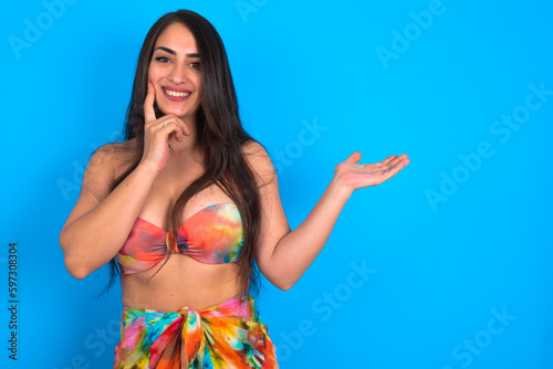 Funny beautiful brunette woman wearing swimwear over blue background hold open palm new product great proposition