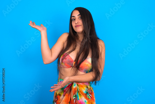 beautiful brunette woman wearing swimwear over blue background pointing aside with both hands showing something strange and saying: I don't know what is this. Advertisement concept.