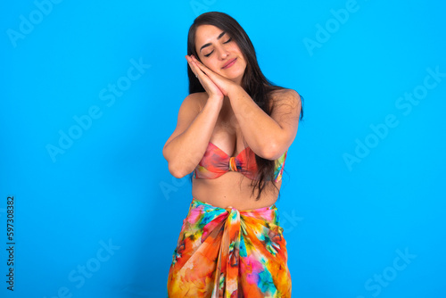 beautiful brunette woman wearing swimwear over blue background sleeping tired dreaming and posing with hands together while smiling with closed eyes.