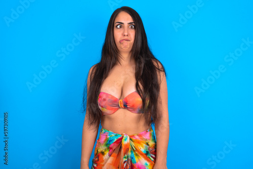 beautiful brunette woman wearing swimwear over blue background making grimace and crazy face, screaming out of control, funny lunatic expressing freedom and wild.