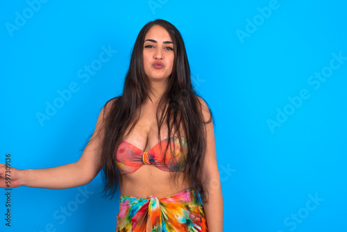 Shot of pleasant looking beautiful brunette woman wearing swimwear over blue background , pouts lips, looks at camera, Human facial expressions