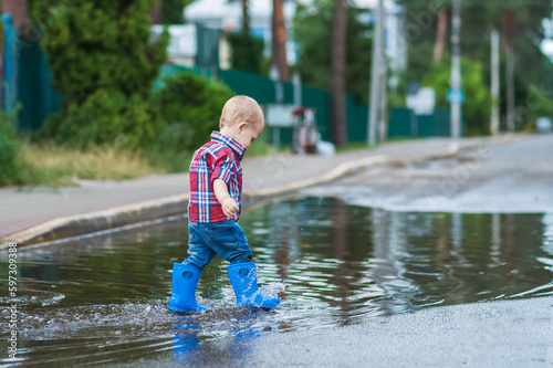 Happy toddler boy running through puddles in rubber boots. Child splashes after rain outside..