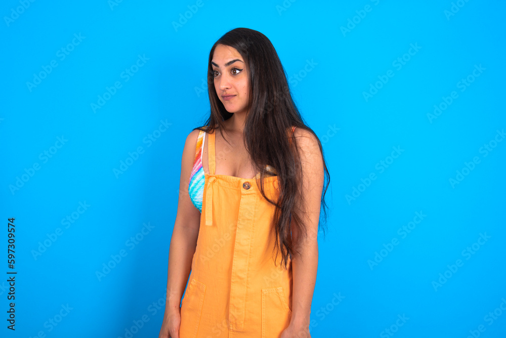 brunette woman wearing orange overalls over blue studio background stares aside with wondered expression has speechless expression. Embarrassed model looks in surprise brunette woman wearing orange overalls over blue studio background stares aside with wondered expression has speechless expression. Embarrassed model looks in surprise