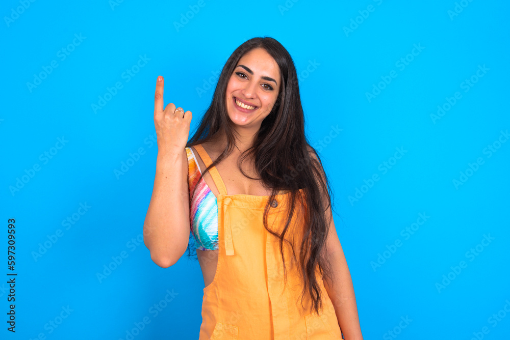 brunette woman wearing orange overalls over blue studio background smiling and looking friendly, showing number one or first with hand forward, counting down brunette woman wearing orange overalls over blue studio background smiling and looking friendly, showing number one or first with hand forward, counting down
