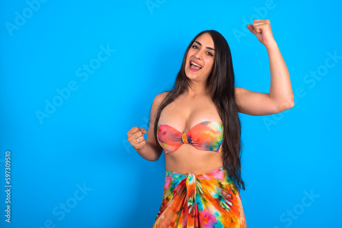 Profile photo of excited beautiful brunette woman wearing swimwear over blue background good mood raise fists screaming rejoicing overjoyed basketball sports fan supporter