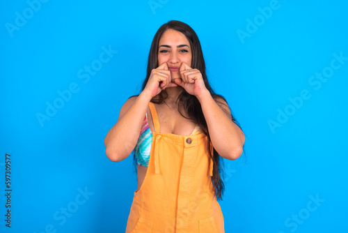 Pleased brunette woman wearing orange overalls over blue studio background with closed eyes keeps hands near cheeks and smiles tenderly imagines something very pleasant