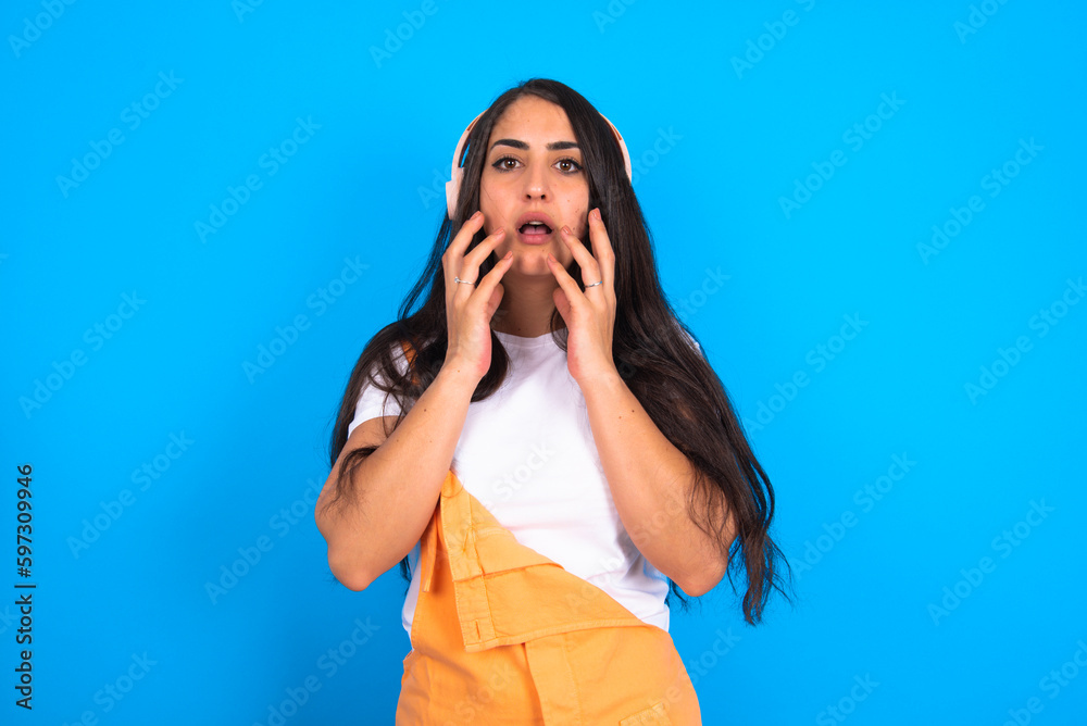 Shocked brunette woman wearing orange overalls over blue studio background stares fearful at camera keeps mouth widely opened wears wireless stereo headphones on ears Shocked brunette woman wearing orange overalls over blue studio background stares fearful at camera keeps mouth widely opened wears wireless stereo headphones on ears
