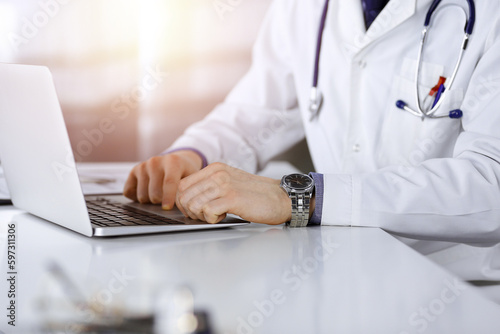 Unknown male doctor sitting and working with laptop in a darkened clinic  glare of light on the background  close-upof hands