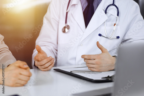 Unknown male doctor and patient woman discussing something while sittingin a darkened clinic, glare of light on the background. Close-up of hands