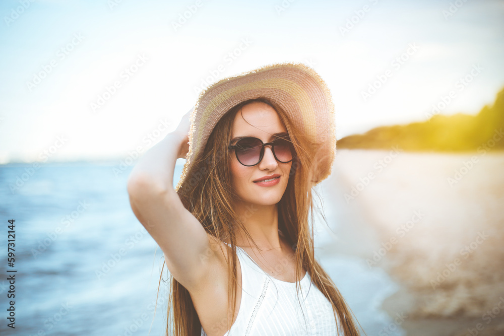 Happy smiling woman in free happiness bliss on ocean beach standing and posing with hat and sunglasses. Portrait of a female model in white summer dress enjoying nature Happy smiling woman in free happiness bliss on ocean beach standing and posing with hat and sunglasses. Portrait of a female model in white summer dress enjoying nature