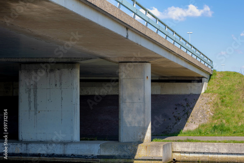 Fototapeta Naklejka Na Ścianę i Meble -  Cement concrete pillar structure of urban bridge crossing over small river, canal or ditch with green grass meadow, Underneath of classic style bridge under blue sky and white clouds, Netherlands.