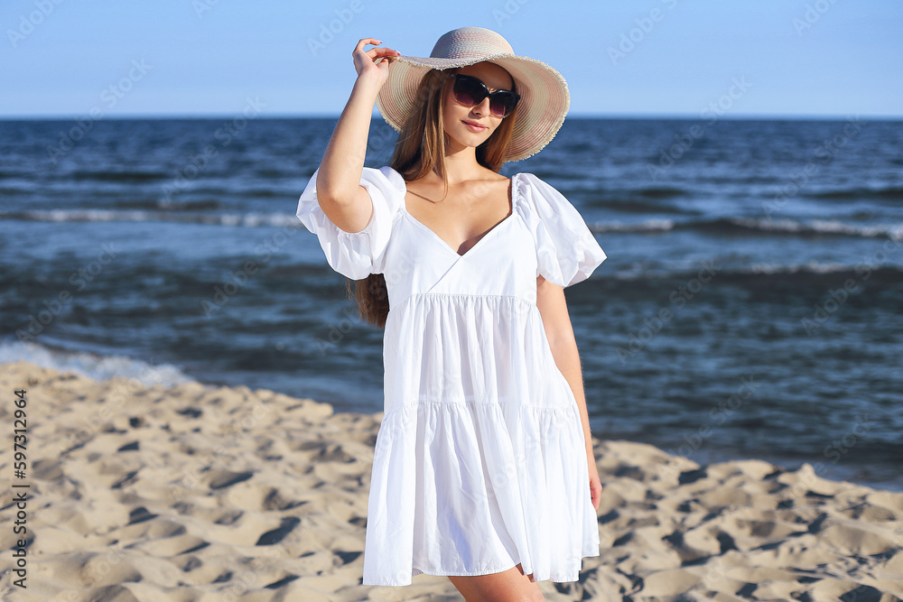 Happy blonde woman is posing on the ocean beach with sunglasses and a hat. Evening sun