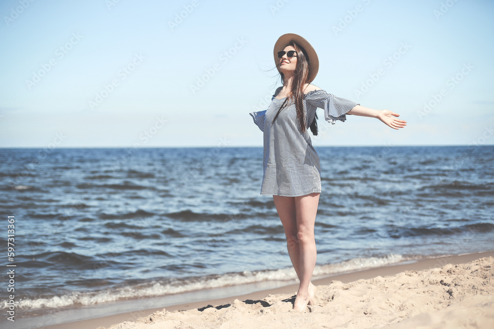 Happy smiling woman in free bliss on ocean beach standing with open hands. Brunette female model in sunglasses and hat enjoying nature during travel holidays vacation outdoors, medium shot