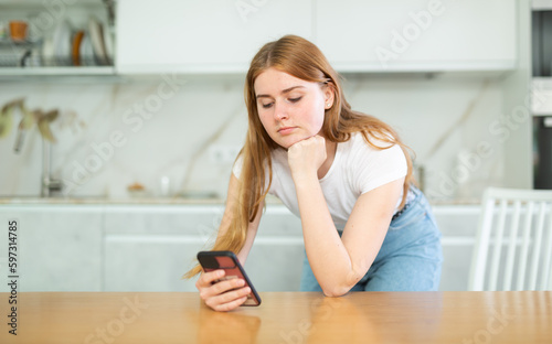 Concentrated European teenage girl in white t-shirt and blue jeans searching info or recipe on smartphone in modern cuisine