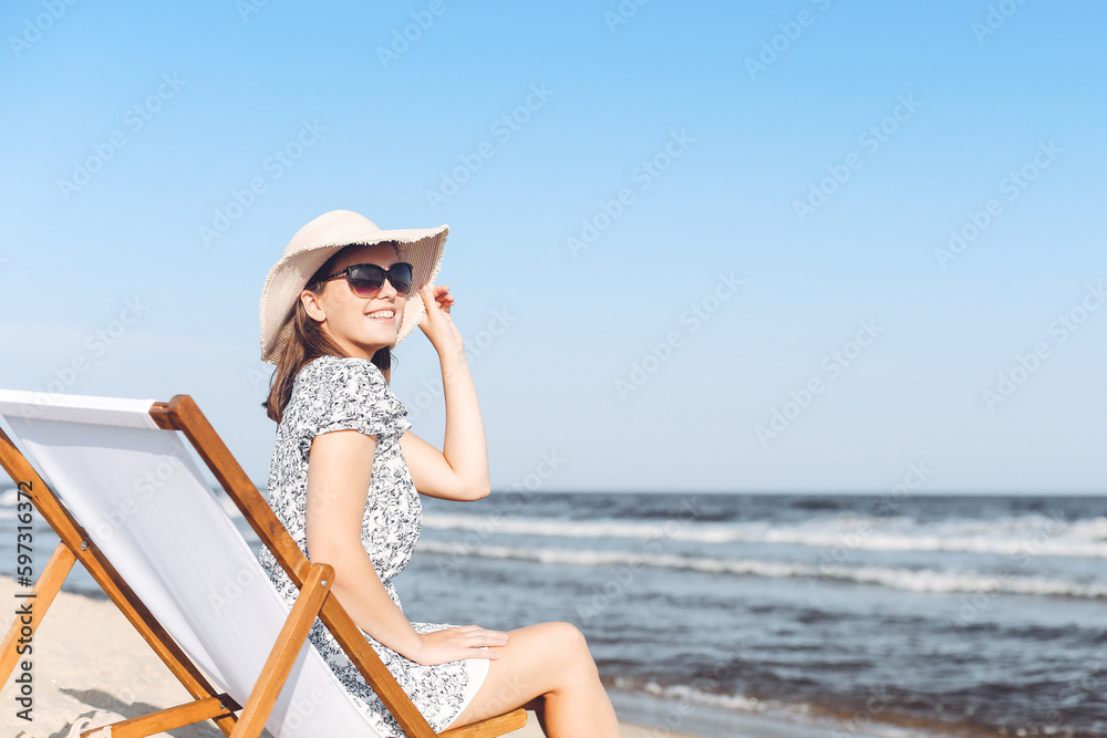 Happy brunette woman wearing sunglasses while relaxing on a wooden deck chair at the ocean beach