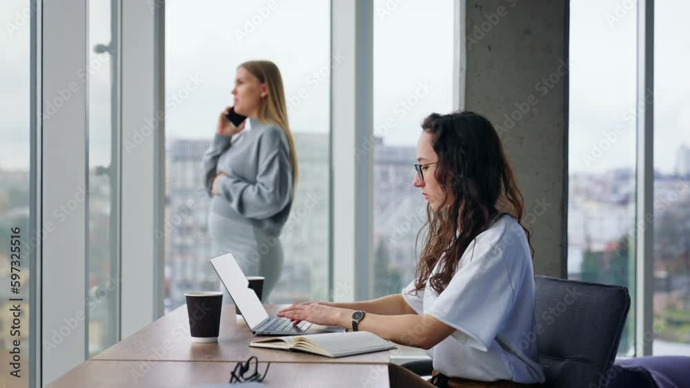Serious focused on work brunette lady works on laptop. Pregnant lady speaks on the phone standing and looking at window at backdrop. Serious focused on work brunette lady works on laptop. Pregnant lady speaks on the phone standing and looking at window at backdrop.