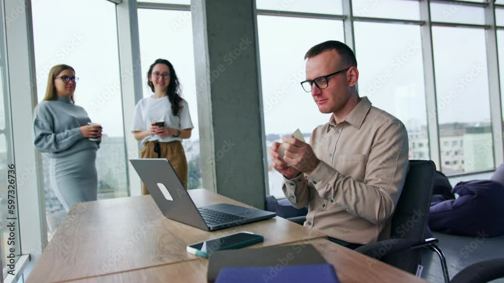 Stressed angry male employee sitting at desk tears a piece of paper. Women standing at backdrop look at male colleague. Blonde women remarks about man’s behavior but he throws the papers around. Stressed angry male employee sitting at desk tears a piece of paper. Women standing at backdrop look at male colleague. Blonde women remarks about man’s behavior but he throws the papers around.
