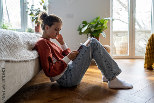 Teens and cyberbullying. Upset teen girl sitting on floor near bed using smartphone at home, scrolling social media. Child spending too much time on phone. Teenagers and gadget addiction photo